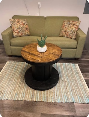 Une table basse ronde en bois, sur laquelle sont posés un vase blanc et un cactus, est placée sur un tapis tissé devant un canapé vert agrémenté de coussins à motifs.