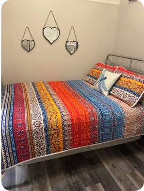 Een bed met een kleurrijk, patroonrijk sprei en twee kussens, een hartvormige spiegel aan de muur en een metalen hoofdbord.