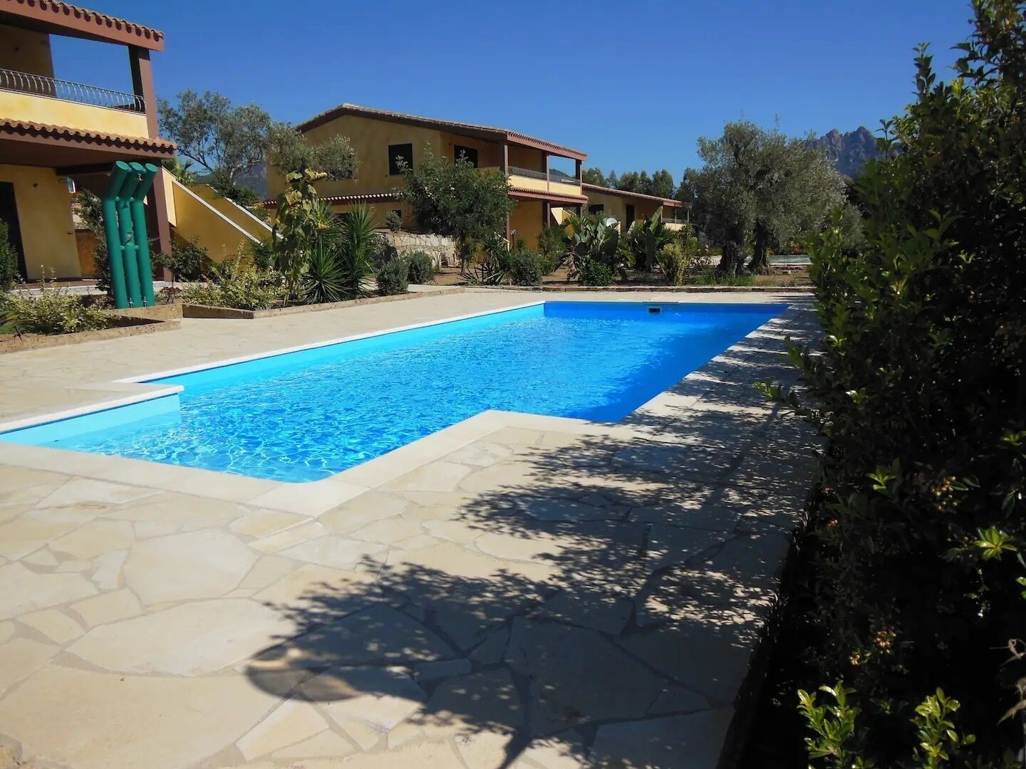 Ein rechteckiger Pool mit einer gefliesten Liegefläche, umgeben von Grünflächen und einem Gebäude mit Balkon.