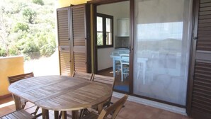 Une terrasse avec une table ronde en bois et des chaises, une porte-fenêtre coulissante donnant sur un espace cuisine, et une vue sur la verdure à l’extérieur.