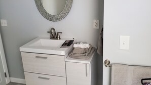 Une salle de bain avec un meuble-lavabo blanc, un miroir rond orné d’un cadre en mosaïque, un porte-serviettes avec une serviette, et un luminaire placé au-dessus du miroir.