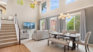 A spacious living room with a staircase, a dining area, and large windows offering views of the outdoors. - Epic 140 inch TV Wall + Game Consoles Galore (Commerce City)