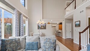 A spacious living room with a large gray sectional sofa, a chandelier, and a view of the backyard through large windows. - Epic 140 inch TV Wall + Game Consoles Galore (Commerce City)