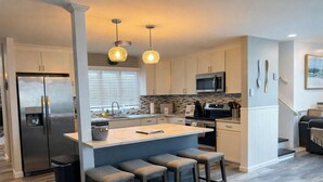 A modern kitchen with a central island, stainless steel appliances, pendant lighting, and a view to the living area.