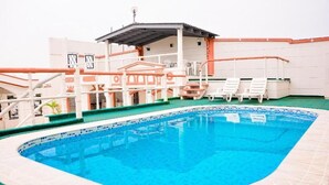 Una piscina con borde de azulejos, una zona de estar techada con sillas y un edificio con tejado de azulejos al fondo.