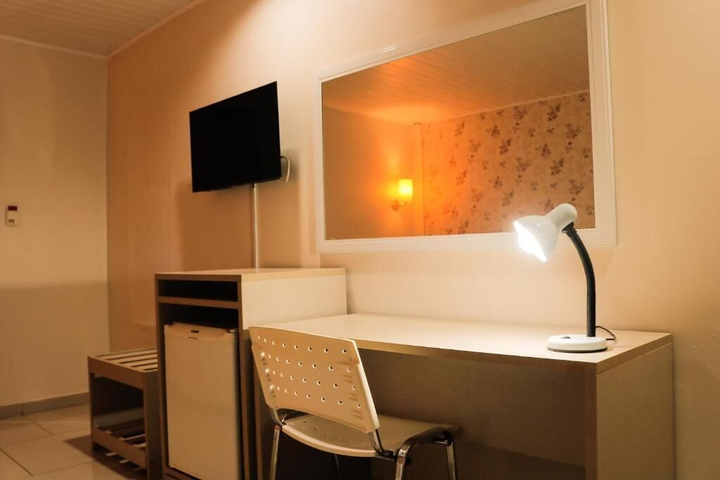 A small, well-lit room with a desk, a chair, a television mounted on the wall, and a window with a light fixture.