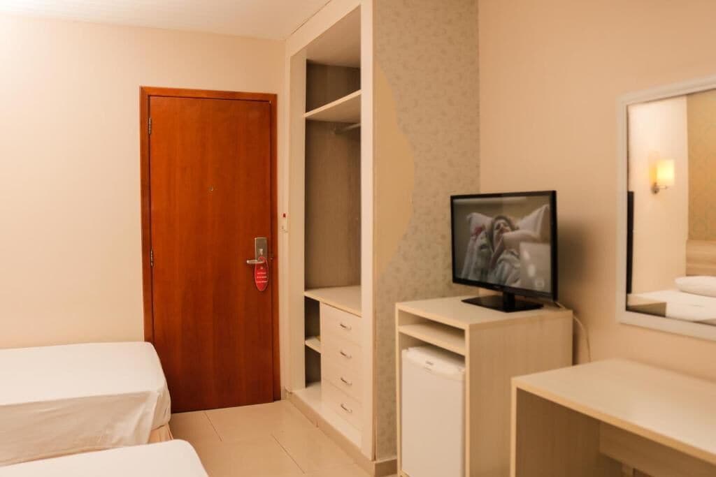 A hotel room with a wooden door, a TV on a desk, a bed, and a wardrobe.
