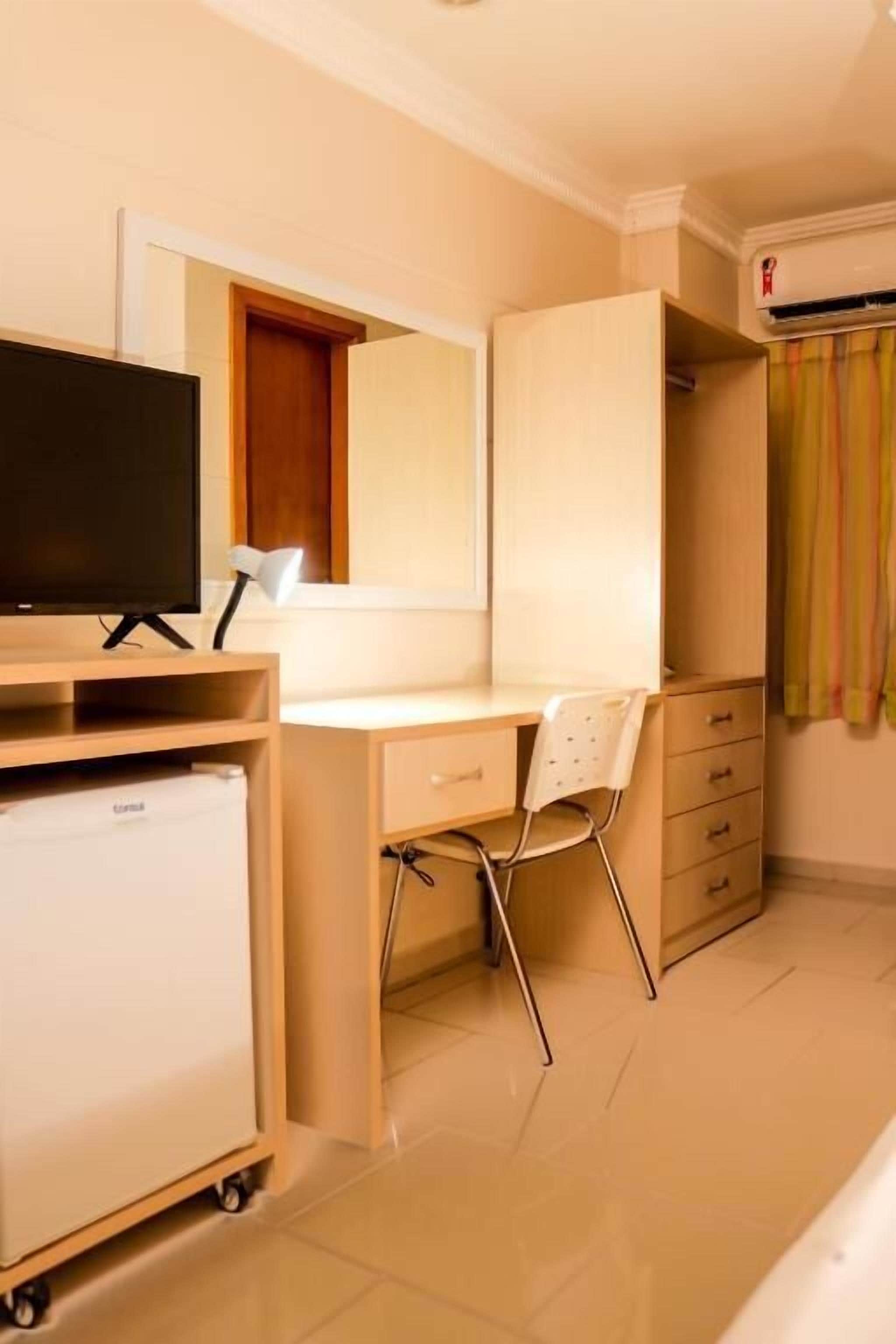 Une chambre d’hôtel de taille réduite, comprenant un lit simple, un bureau avec une chaise, une télévision, un mini-réfrigérateur et une armoire.