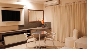 Habitación de hotel con televisión de pantalla plana en la pared, una mesa redonda con dos sillas, cama y una ventana grande con cortinas.