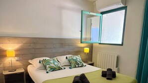 A hotel room with a wooden headboard, a bed with a green bedspread, two bedside lamps, a mirror, and a radiator. - Cozy villa 🌞, air-conditioned, 5' walk to San Ciprianu beach 🌊 (Lecci)