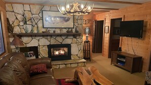 Un salon chaleureux avec une cheminée en pierre, un canapé en cuir, une table basse en bois et un téléviseur fixé au mur.