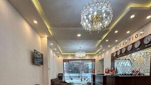 A hotel reception area with a patterned sofa, a chandelier, a reception desk with clocks, and a view of the outside. - Hotel Diyor (Samarkand)