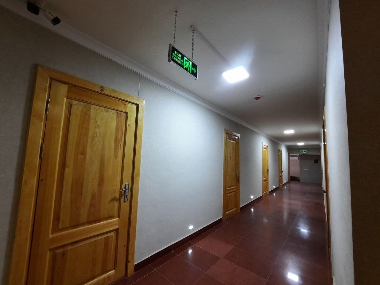A long, narrow hallway with wooden doors on one side and a sign indicating an exit.