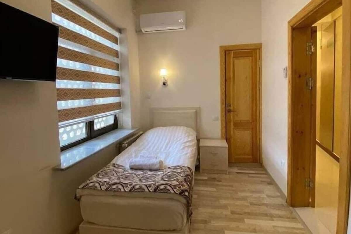 Una habitación pequeña y estrecha con una cama, una ventana con persianas, un televisor montado en la pared, aire acondicionado y una puerta de madera.