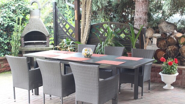 Eine Terrasse im Freien mit einem Steinpizzaofen, Korbstühlen und einem Tisch mit roten Unterlayscheiben.