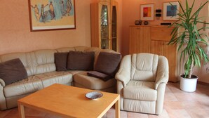 Ein Wohnzimmer mit Sofa, Sessel und Couchtisch. Ein Schrank mit Glastüren und ein Topfpflanze sind ebenfalls vorhanden.