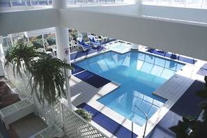 Una piscina interior con baldosas azules, sillas de descanso y palmeras.