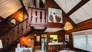 Interior acogedor de una cabaña con escalera, paredes de madera, zona de comedor con sillas azules y cocina con refrigerador.