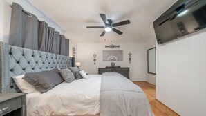 A bedroom with a tufted headboard, a bed with white and gray bedding, a ceiling fan, a TV mounted on the wall, and a dresser with a mirror.