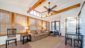 A spacious living room with a large sofa, a coffee table, and a dining area with a table and chairs. There is a ceiling fan and large windows with blinds.