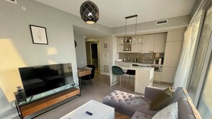 A modern living room with a flat-screen TV, a white coffee table, and a tufted sofa. A kitchen area with white cabinets and a dining table is visible in the background.