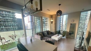 A modern living space with a sofa, coffee table, and dining area. Large windows offer views of a cityscape.