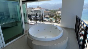 Un balcón con jacuzzi, puertas de cristal y vistas a montañas y una playa.