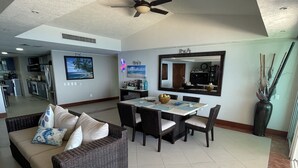 Amplia sala de estar con mesa de comedor y mobiliario de mimbre oscuro, ventilador de techo y cocina al fondo.