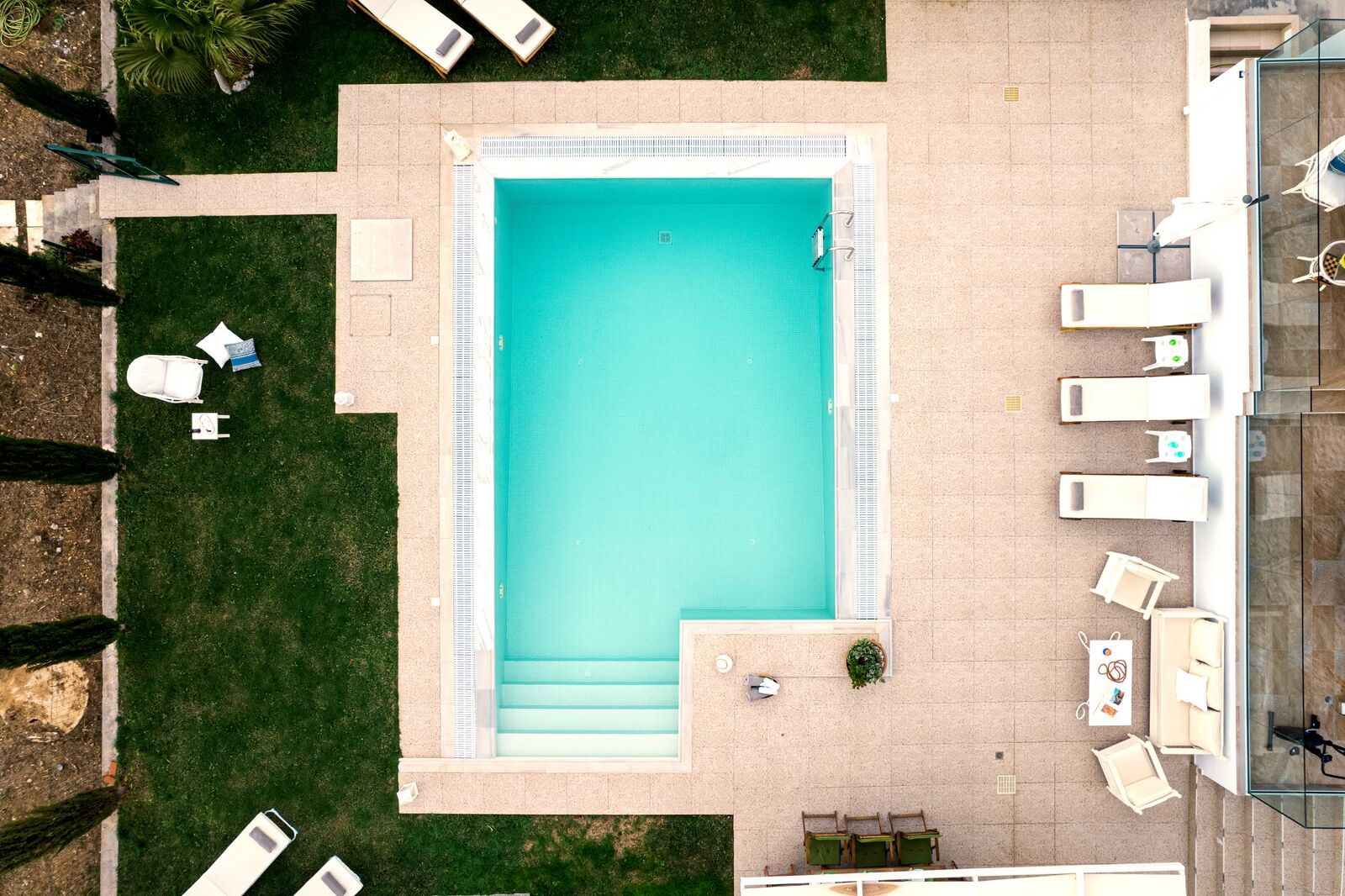 Une vue aérienne d’une piscine rectangulaire, entourée d’une terrasse carrelée, avec quelques chaises longues et un petit coin salon comprenant une table et des chaises.