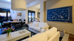 A modern living room with a dining area, a large window, and a blue wall with a nautical-themed poster. - Smaris Collection luxury villas.Sea side villa 5 min.from the beach ! (Σίσι)