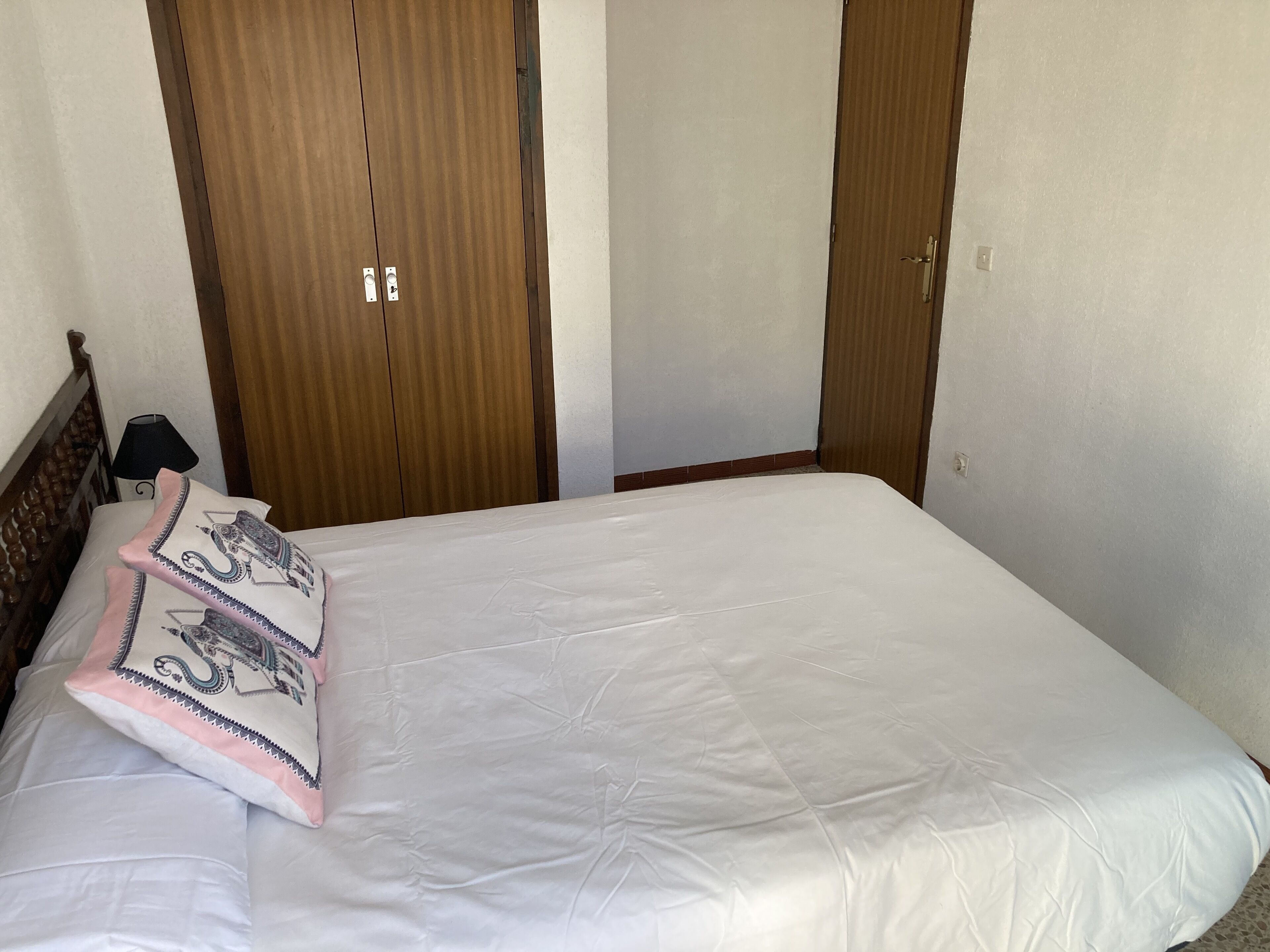 Cama bem arrumada com lençóis brancos e almofadas decorativas, cabeceira de madeira e guarda-roupa de madeira com espelho.