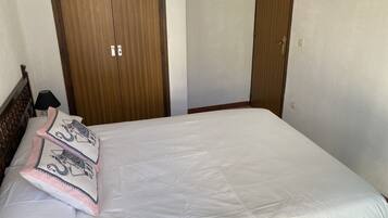 Cama bem arrumada com lençóis brancos e almofadas decorativas, cabeceira de madeira e guarda-roupa de madeira com espelho.