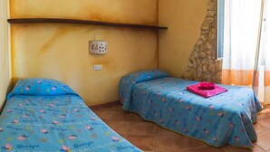 A bedroom with two beds, each with a blue bedspread featuring cartoon characters and the name 'George'. There is a stone wall and a window with curtains.