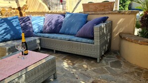 A wicker sofa with blue and purple cushions, a glass table with a bottle and glass, and a stone floor.