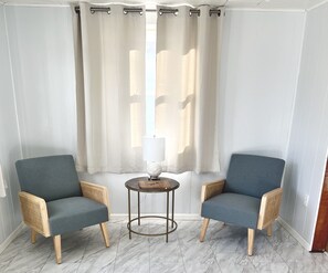 Une pièce d’angle avec deux fauteuils gris, une petite table ronde avec une lampe et un rideau blanc.
