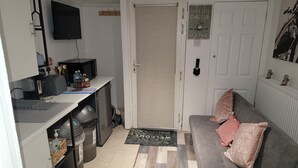 Un espacio habitable compacto con kitchenette, un sofá gris con cojines decorativos, un televisor montado en la pared y una puerta con un letrero que dice “TAKE LIFE ONE CUP AT A TIME”.