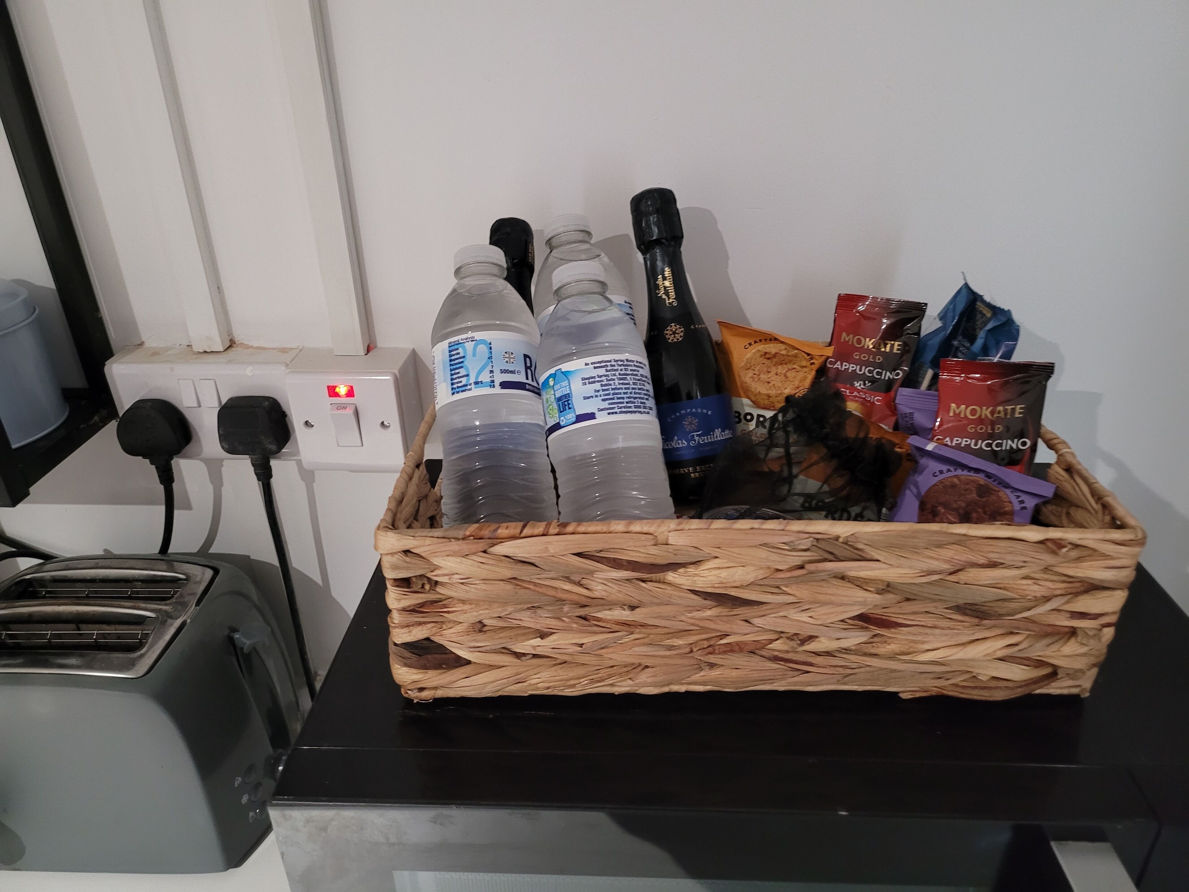 A basket containing bottled water, a bottle of champagne, and coffee pods on a table next to a power outlet and a toaster.