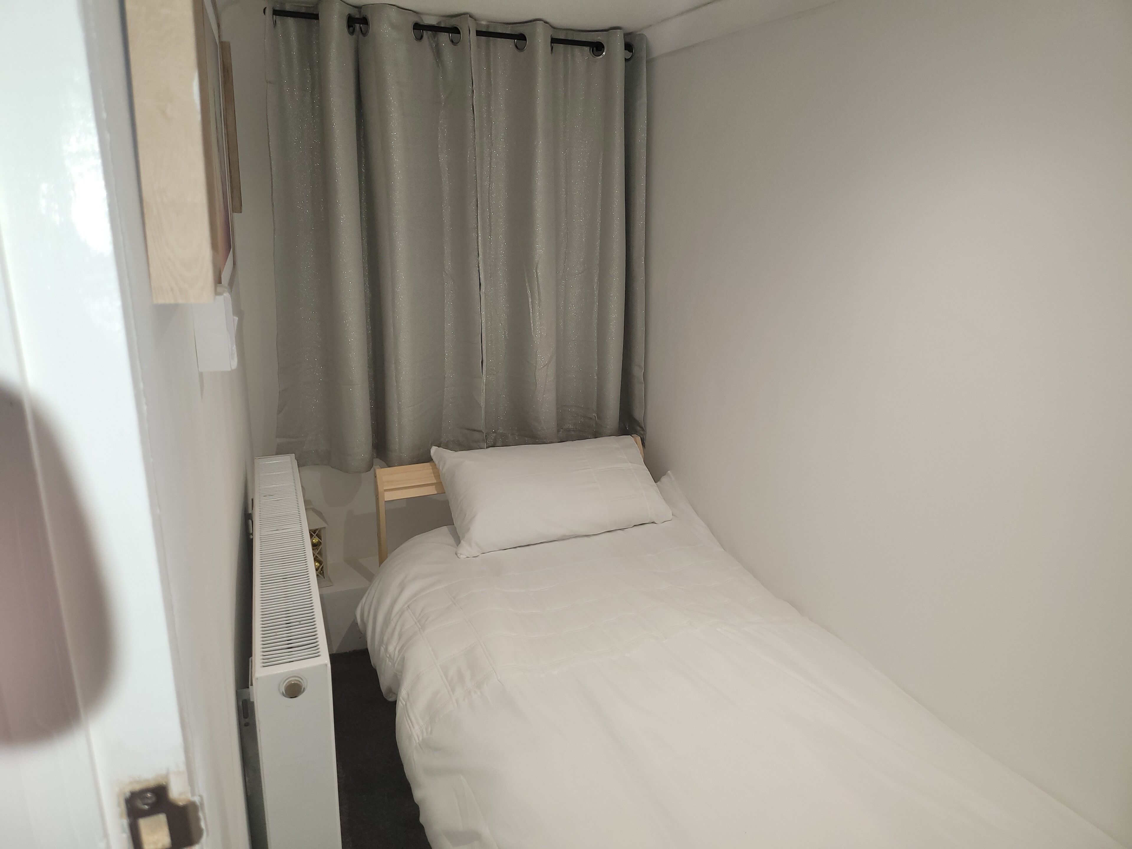 A small, single-bed room with a white bed, a wooden nightstand, a white radiator, and grey curtains.