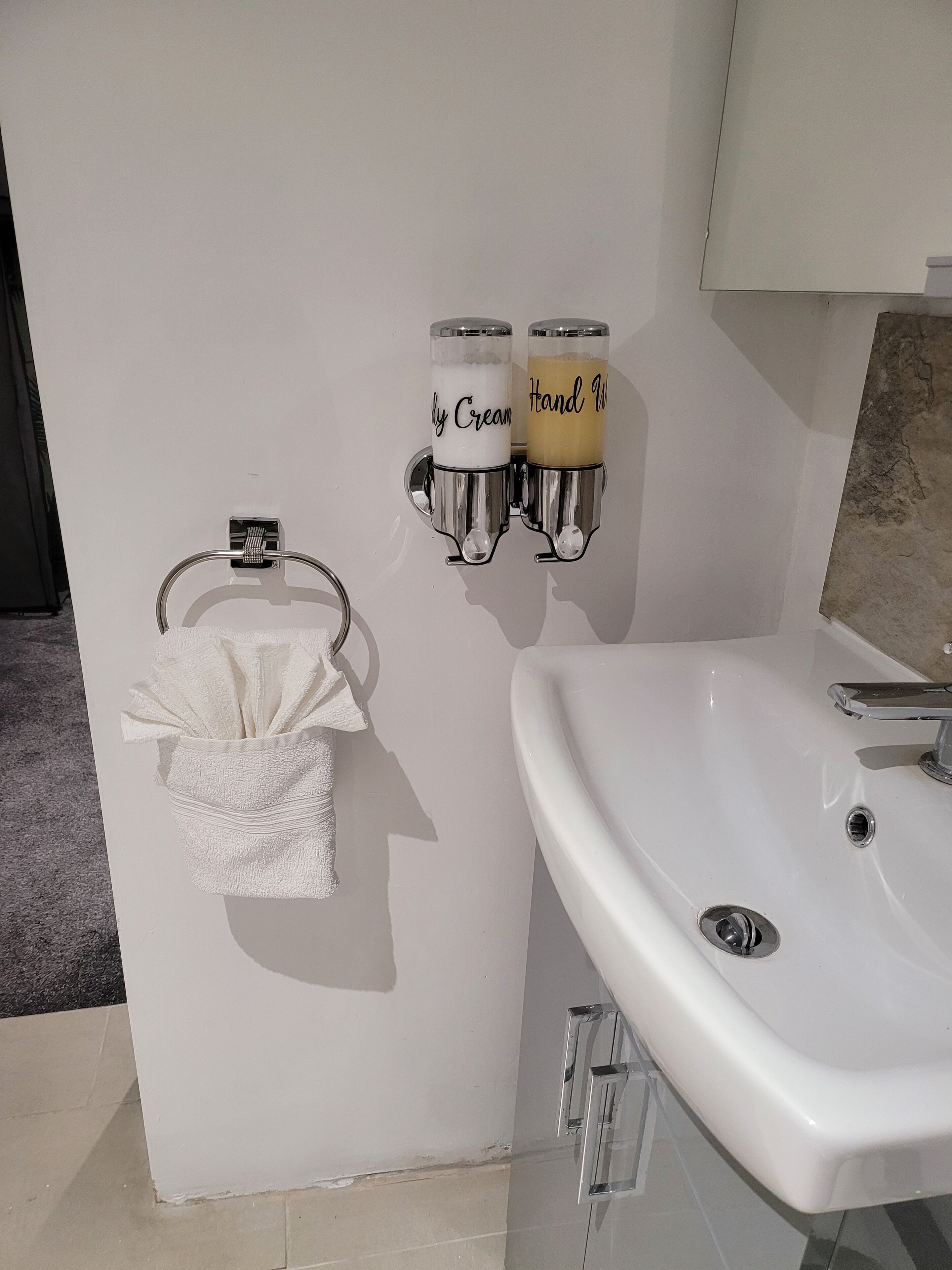 Et badeværelse med en hvid vask, et håndklædestativ med et hvidt håndklæde og to sæbedispensere mærket 'Daily Cream' og 'Hand Wash'.