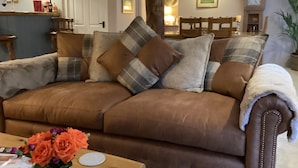 A brown leather sofa with plaid cushions, a wooden coffee table with a vase of orange roses, and a wooden chair in the background. - The Snuggery a Cosy Country Cottage (Wellington)