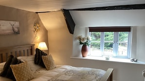 A bedroom with a bed, pillows, a lamp, a vase with flowers, and a window with a view of greenery. - The Snuggery a Cosy Country Cottage (Wellington)