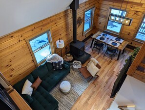 Indretning i en hyggelig hytte med sofa i grøn farve, spisebord og brændeovn.