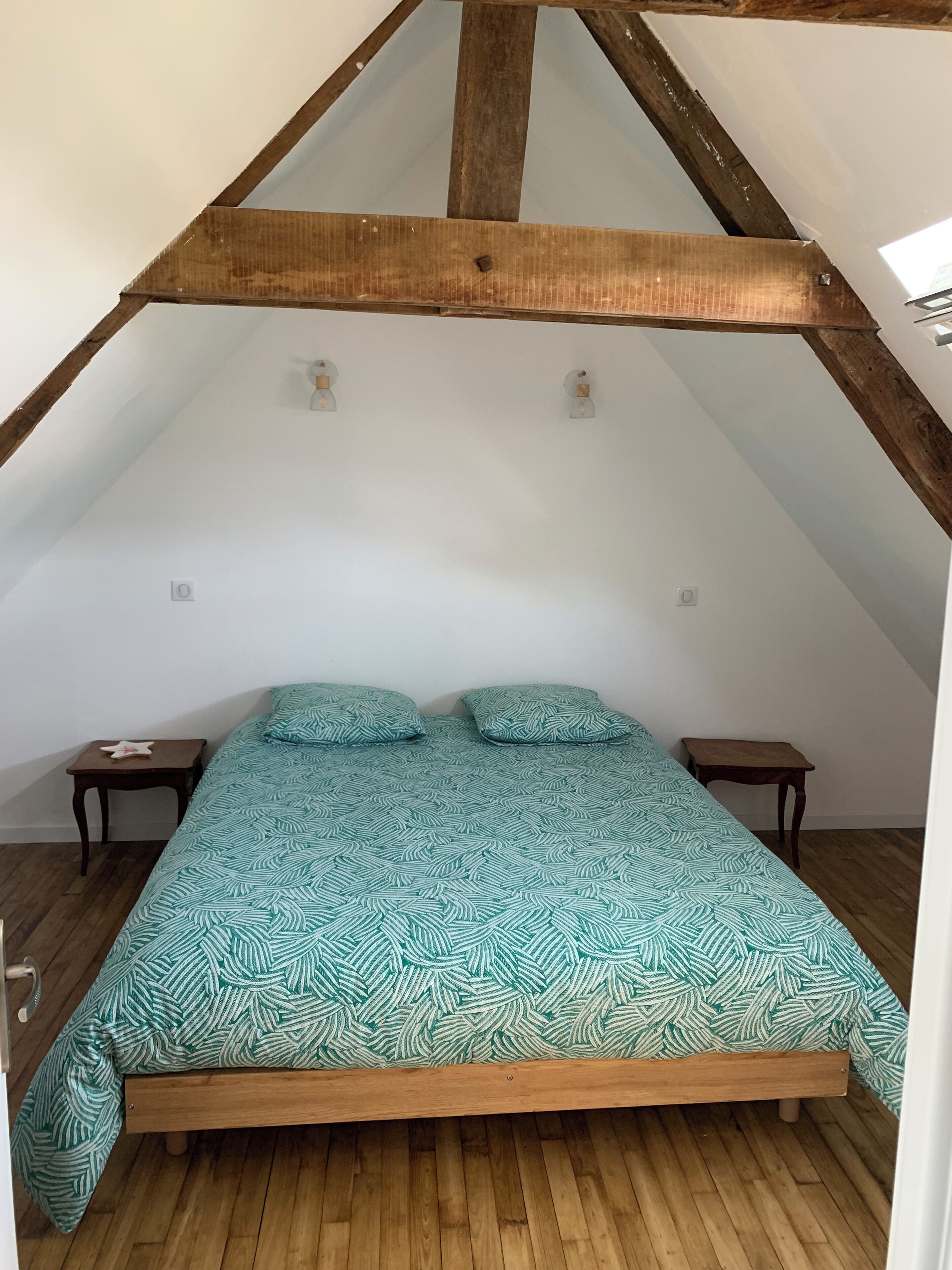 Un dormitorio con techo inclinado, una cama con ropa de cama estampada, dos mesitas de noche y suelo de madera.