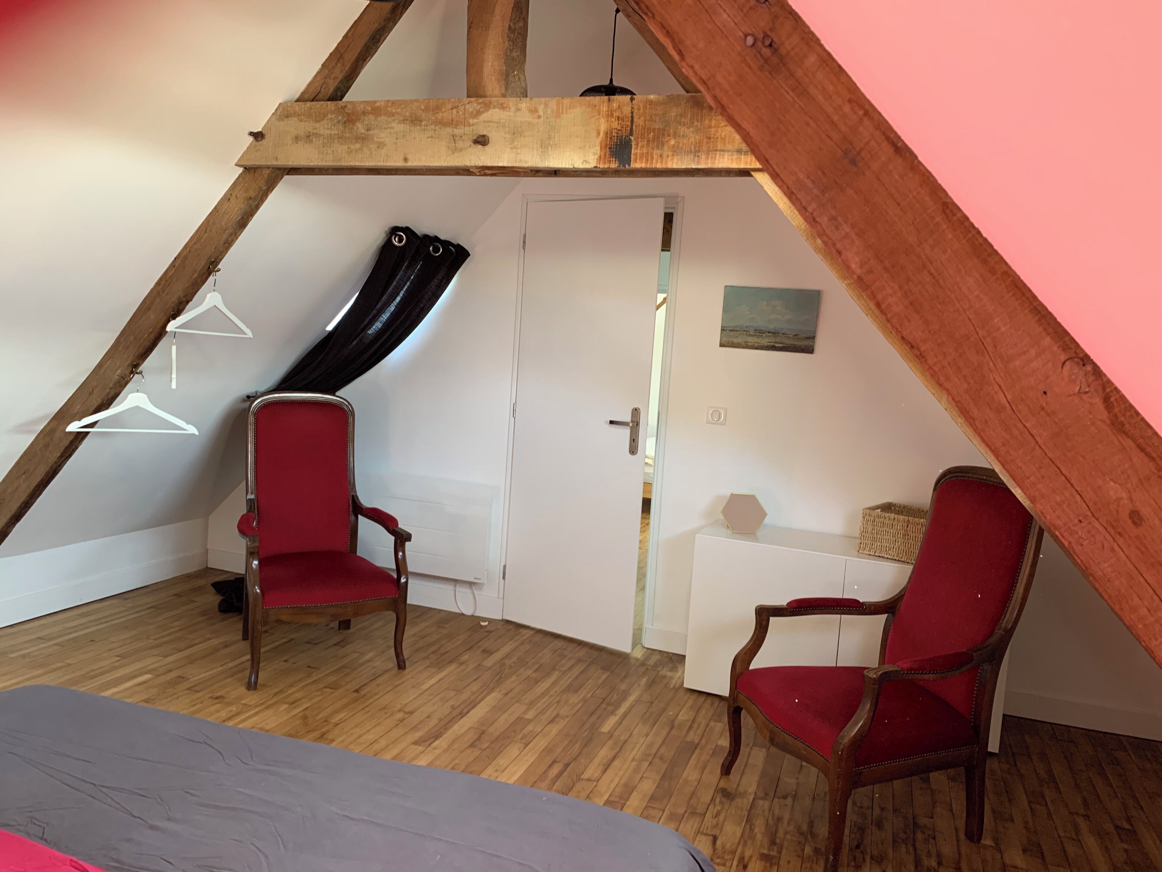 Une pièce avec un plafond en pente, des poutres en bois, deux chaises rouges, une porte blanche et un petit meuble blanc.