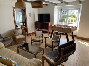A living room with a sofa, armchairs, a coffee table, a foosball table, a television, and a window with a view of greenery.