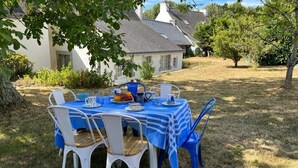 Un espace de restauration en plein air avec une table dressée pour quatre personnes, entourée de chaises blanches, sous un arbre.