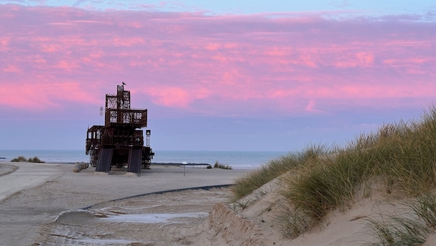 Une plage avec une grande structure métallique, peut-être un phare, et des dunes de sable.