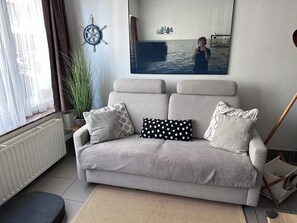 Un salon avec un canapé gris, une plante en pot, un radiateur et un tableau représentant une personne sur un bateau.