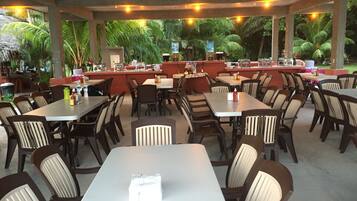 A restaurant with multiple tables and chairs, a counter with food and beverages, and a view of tropical plants.