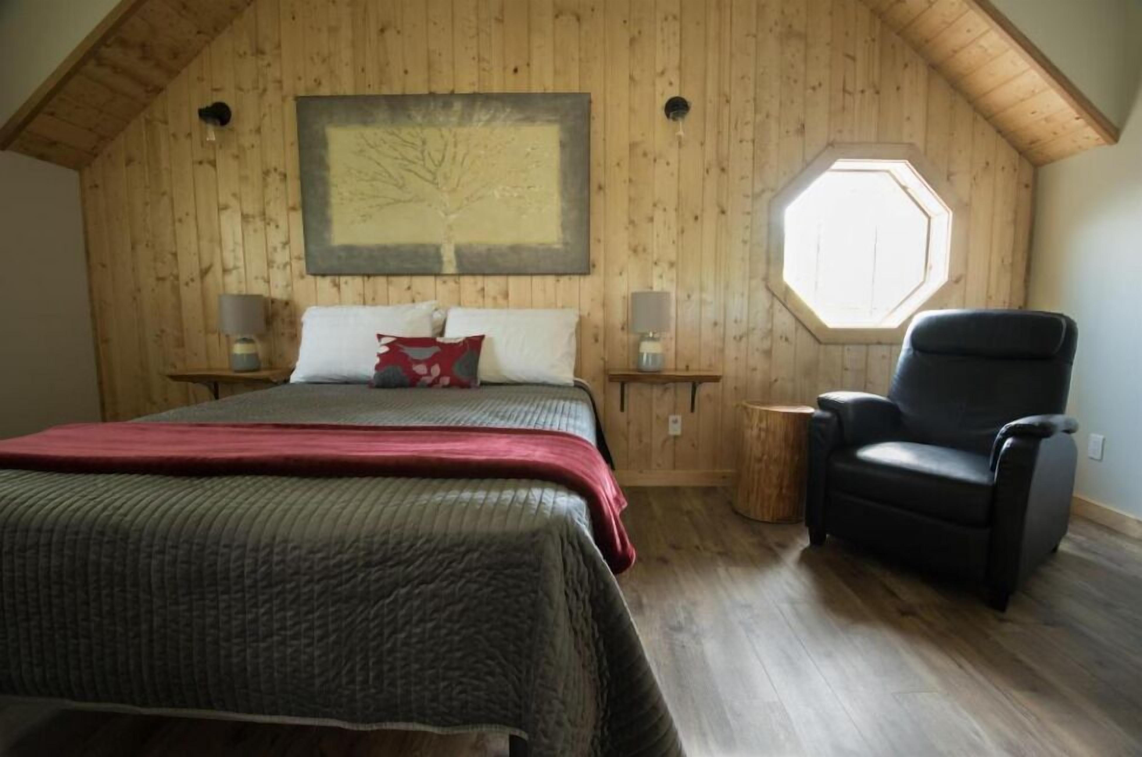 A bedroom with a wooden ceiling, a large bed with a red blanket, a black chair, a window, and a painting on the wall.